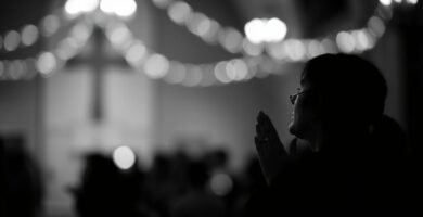 Schwarz-Weiß-Bild einer betenden Frau in einer Kirche mit farbigen Lichtern an der Decke und einem Kreuz auf dem Sockel im Hintergrund, der Hintergrund ist unscharf