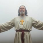 Bild eines Priesters in weißem Habit mit einem Kreuz auf der Brust, die Arme ausgestreckt, im Hintergrund weißer Nebel.