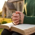 Nahaufnahmen einer Frau, die mit einem Rosenkranz in der Hand auf einer Bank sitzt und in der Bibel liest; ihr Gesicht ist nicht zu sehen. Im Hintergrund befindet sich eine Pfarrkirche inmitten eines Feldes.