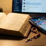 Nahaufnahme einer aufgeschlagenen Bibel neben einem Mobiltelefon mit Social-Media-Symbolen auf dem Bildschirm und einem Rosenkranz daneben. Im Hintergrund ein Laptop mit einer Social-Media-Konversation.